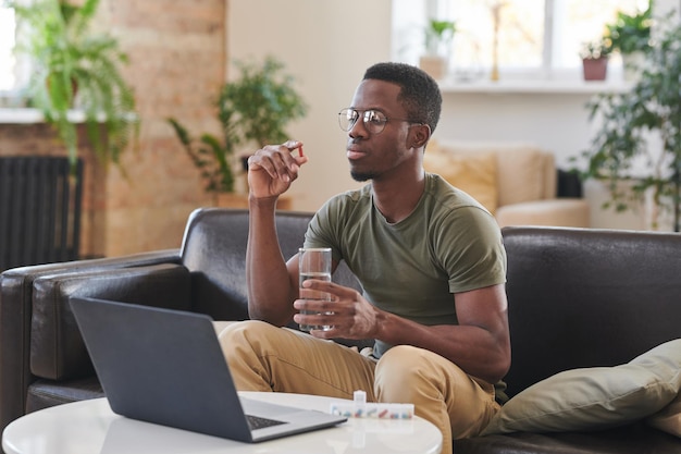 Giovane adulto afroamericano che indossa occhiali da vista seduto sul divano davanti al computer portatile che tiene il vetro