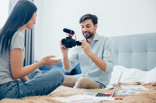 Giovane adulto allegro che registra la sua ragazza su una macchina fotografica mentre era seduto su un letto con grafici