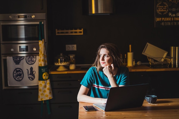 Giovane adulto caucasico seduto nella sua cucina con il suo laptop che lavora da casa guardando lontano.