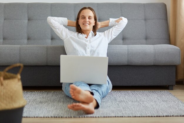 Giovane adulto felice donna sicura di sé che indossa camicia bianca e jeans seduto con il taccuino sulle gambe, braccia alzate dietro la testa, guardando la telecamera, libero professionista soddisfatto del lavoro finito.