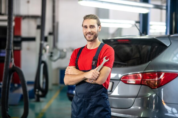 Giovane adulto fiducioso uomo in uniforme da lavoro in piedi vicino a automobili in autosalone