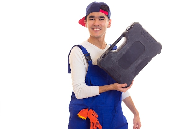 Giovane affascinante con capelli neri in camicia bianca e tuta blu con guanti arancioni e snapback blu che tiene la cassetta degli attrezzi su sfondo bianco in studio