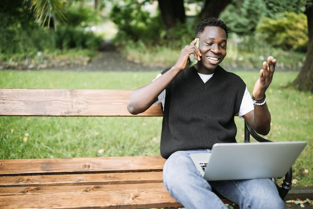 Giovane africano che parla al telefono lavorando al computer portatile nel parco felice soddisfatto