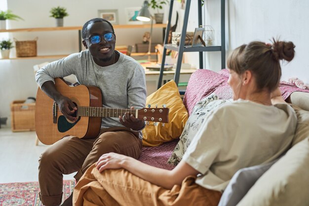 Giovane africano che suona la chitarra e canta canzoni per la sua ragazza sul divano della stanza
