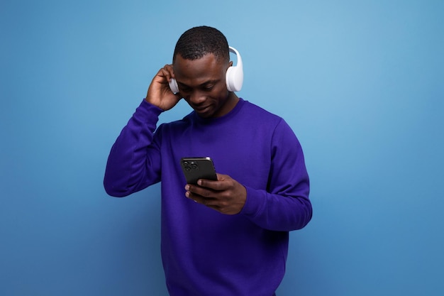 Giovane africano in maglione blu con cuffie bluetooth senza fili che guardano video sul telefono