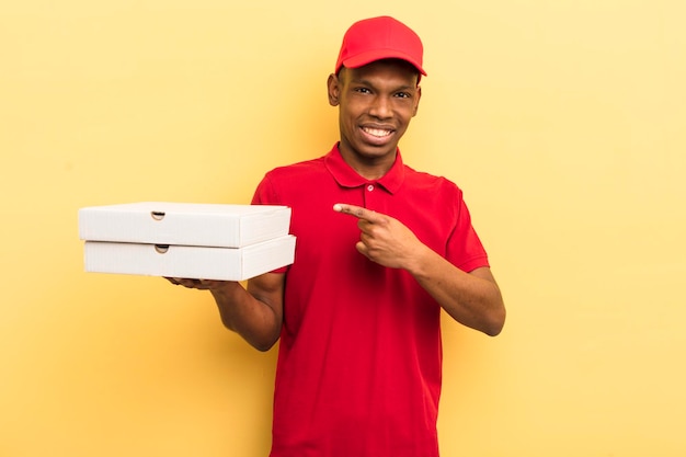 Giovane afro nero che sorride allegramente sentendosi felice e indicando il concetto di uomo di consegna della pizza laterale