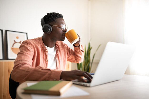 Giovane afroamericano che beve caffè, ascolta musica e lavora con il portatile.