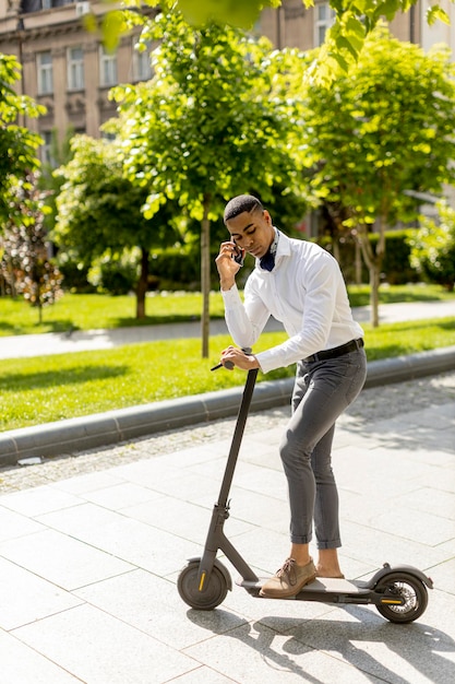 Giovane afroamericano che usa il telefono cellulare mentre è in piedi con uno scooter elettrico su una strada