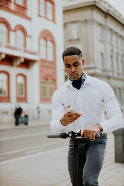 Giovane afroamericano che usa il telefono cellulare mentre è in piedi con uno scooter elettrico su una strada