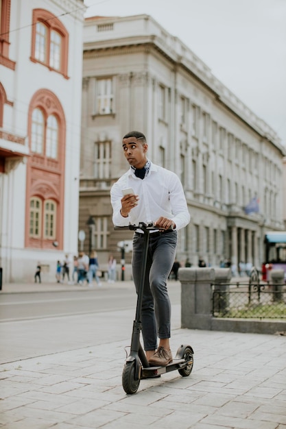 Giovane afroamericano che usa il telefono cellulare mentre è in piedi con uno scooter elettrico su una strada