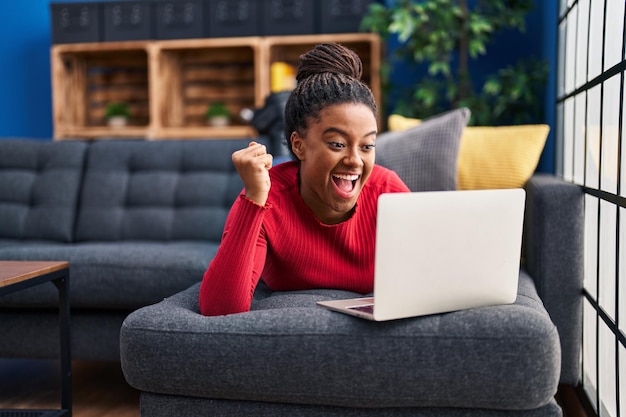 Giovane afroamericano con le trecce che lavora utilizzando il computer portatile urlando orgoglioso, celebrando la vittoria e il successo molto eccitato con le braccia alzate