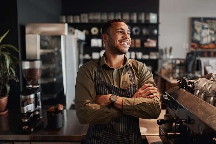 Foto giovane afroamericano proprietario di una piccola caffetteria in piedi dietro il bancone indossando un grembiule con le braccia incrociate guardando lontano
