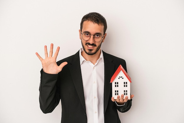 Giovane agente immobiliare uomo in possesso di un modello di casa isolata su sfondo bianco sorridente allegro che mostra il numero cinque con le dita