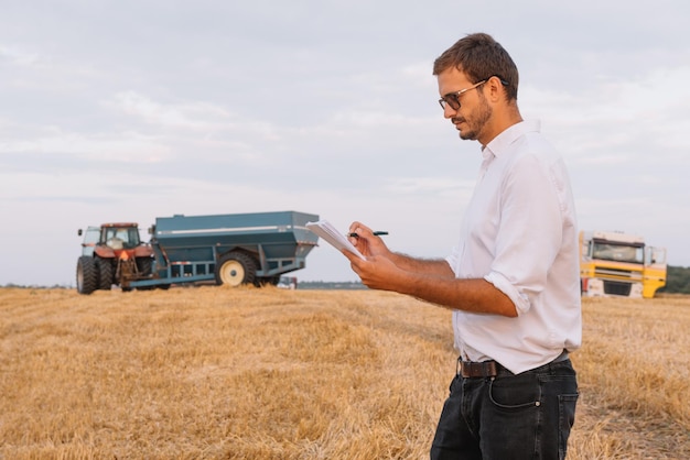 Giovane agricoltore attraente con laptop in piedi nel campo di grano con mietitrebbia in background