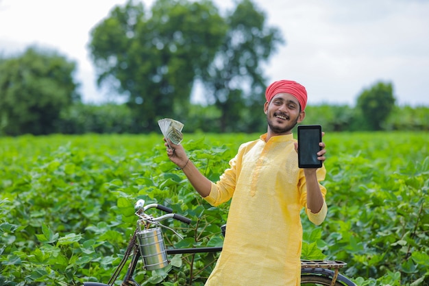 Giovane agricoltore indiano che mostra smartphone e soldi al campo di cotone