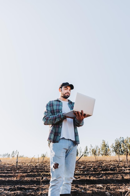 Giovane agricoltore latino che lavora in telelavoro sul suo laptop nel mezzo del suo terreno agricolo Agricolo