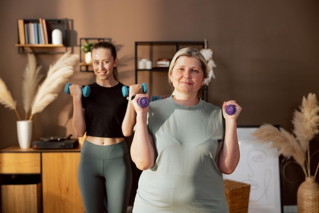 Giovane aiutante che si riscalda facendo sport con una donna anziana che tiene in braccio manubri colorati