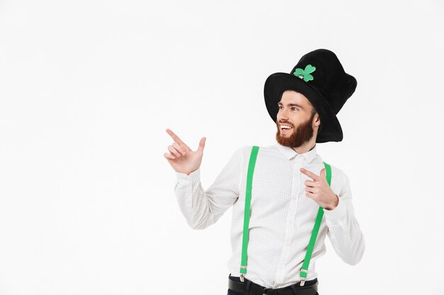 Giovane allegro che celebra il giorno di StPatrick isolato sopra il muro bianco, dito puntato