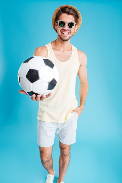 Giovane allegro con cappello e occhiali da sole che ti dà una palla da calcio sul muro blu