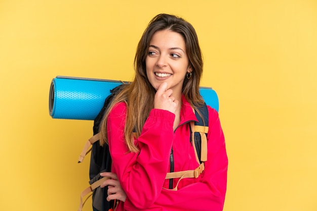 Giovane alpinista con un grande zaino isolato su sfondo giallo guardando di lato e sorridente