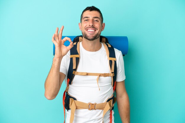 Giovane alpinista uomo caucasico con un grande zaino isolato su sfondo blu che mostra segno ok con le dita
