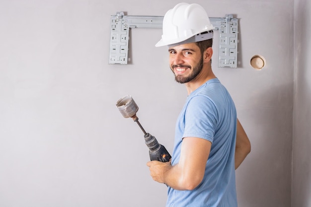 Giovane appaltatore che tiene un trapano con sega a tazza per perforare il muro e installare un condizionatore d'aria sorridente guardando la fotocamera