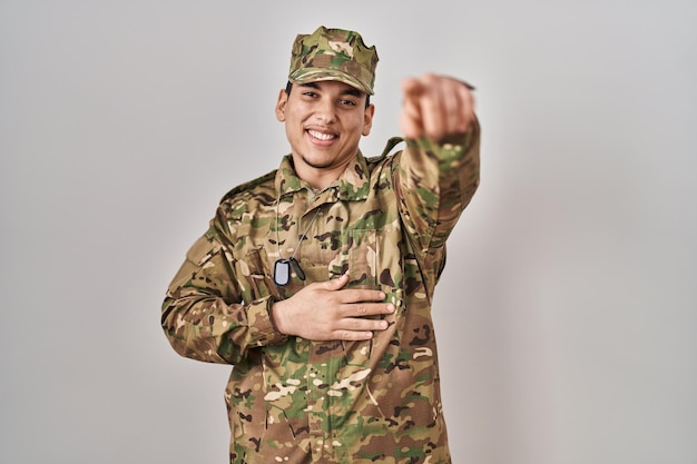 Giovane arabo che indossa l'uniforme dell'esercito mimetico che ride di te, puntando il dito verso la telecamera con la mano sul corpo, espressione di vergogna