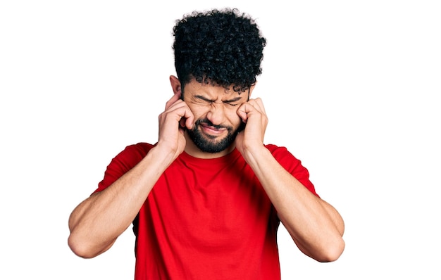 Giovane arabo con la barba che indossa una maglietta rossa casuale che copre le orecchie con le dita con un'espressione infastidita per il rumore della musica forte concetto sordo