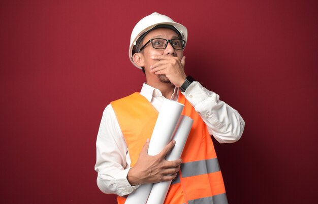 Giovane architetto asiatico che indossa il casco e tiene in mano un piano di carta del progetto con la bocca che copre la mano.