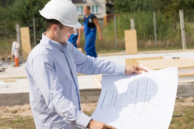 Giovane architetto che controlla un disegno strutturale o un progetto mentre si trova sul cantiere di una nuova casa