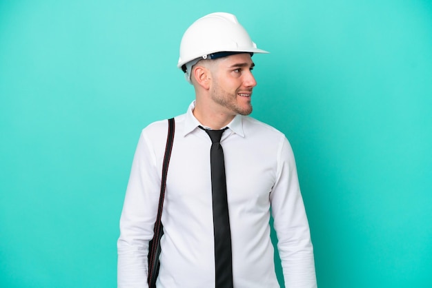 Giovane architetto uomo caucasico con casco e azienda progetti isolati su sfondo blu guardando al lato e sorridente