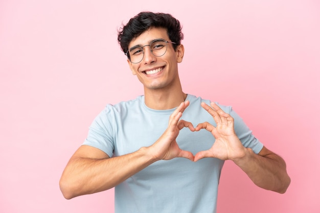 Giovane argentino isolato su sfondo rosa Con gli occhiali che fanno il cuore con le mani