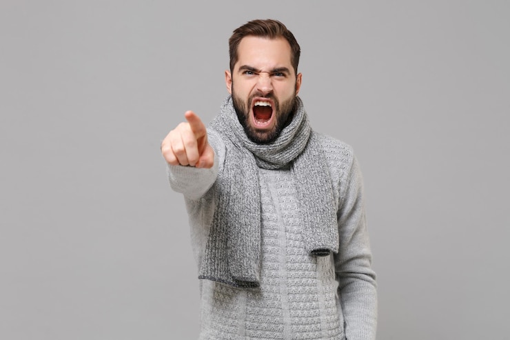 Foto giovane arrabbiato in maglione grigio sciarpa in posa isolato su sfondo grigio in studio. stile di vita sano della moda, concetto di stagione fredda. simula lo spazio della copia. punta il dito indice sulla telecamera urlando imprecazioni.
