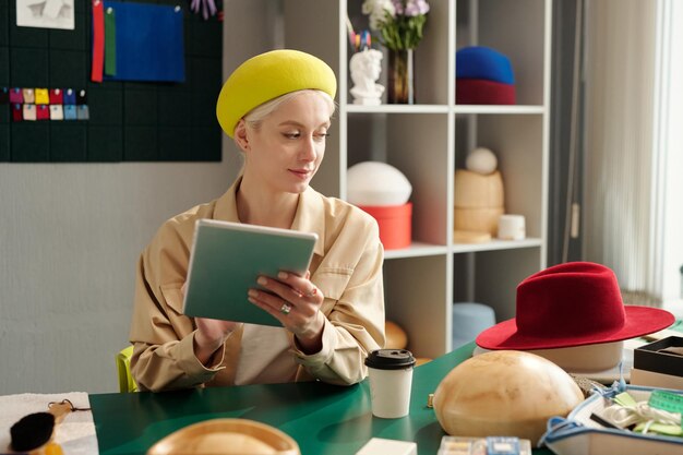 Giovane artigiana con tablet guardando il nuovo cappello cremisi in feltro