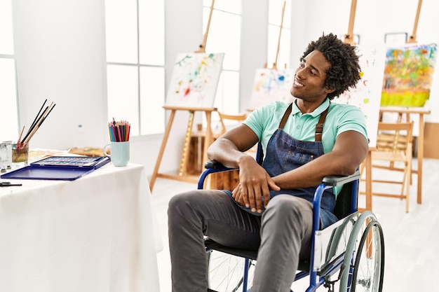 Giovane artista afroamericano seduto sulla sedia a rotelle presso lo studio d'arte che distoglie lo sguardo a lato con il sorriso sull'espressione naturale del viso che ride fiducioso