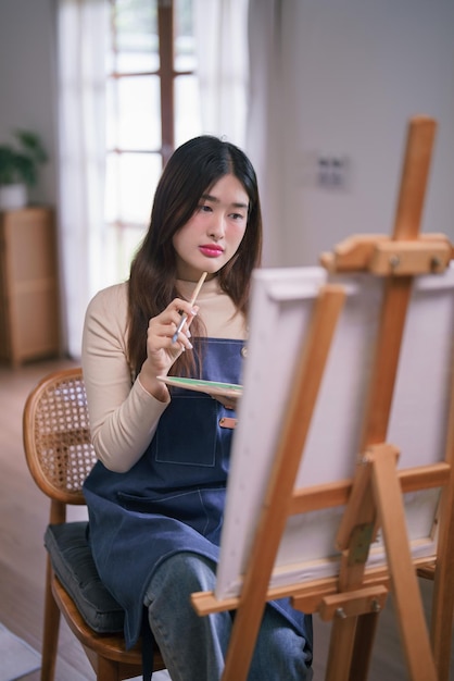Giovane artista asiatica seduta di fronte a una tela e pensando all'idea di disegnare e dipingere opere d'arte