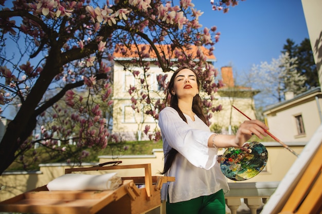 Giovane artista della donna del brunette che tiene in mani una spazzola e una gamma di colori.