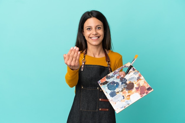 Giovane artista donna caucasica che tiene una tavolozza isolata su sfondo blu che invita a venire con la mano. Felice che tu sia venuto