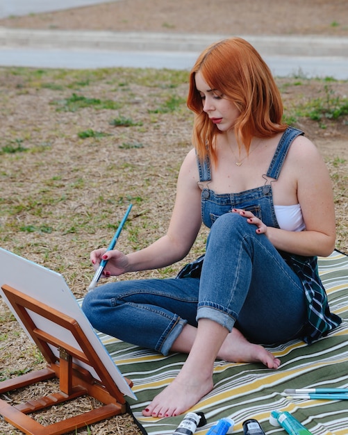 Giovane artista femminile caucasica rossa dipinge una tela su un piccolo cavalletto con un pennello nel parco all'aperto