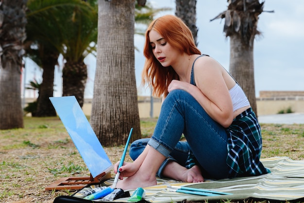 Giovane artista femminile caucasica rossa dipinge una tela su un piccolo cavalletto con un pennello nel parco all'aperto