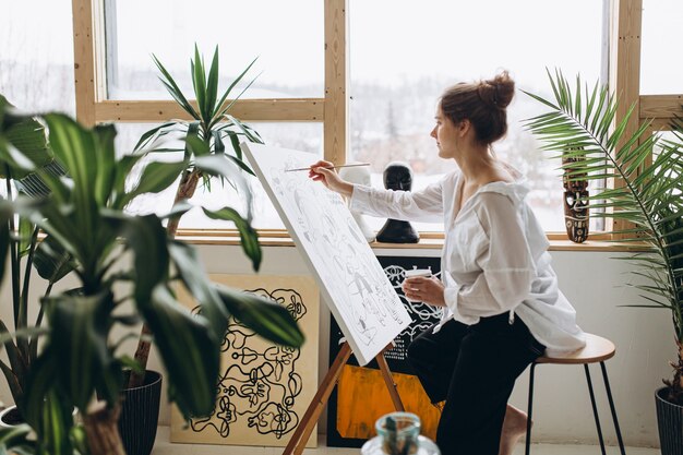 Giovane artista femminile in camicia bianca e pantaloni neri che disegna un'immagine astratta sul cavalletto