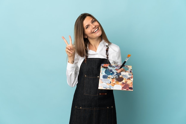 Giovane artista slovacco donna isolata sulla parete blu sorridendo e mostrando il segno di vittoria