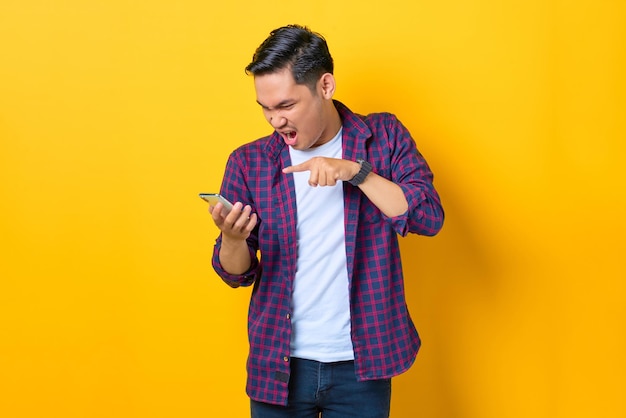Giovane asiatico arrabbiato in camicia a quadri usando un telefono cellulare e urlando isolato su sfondo giallo