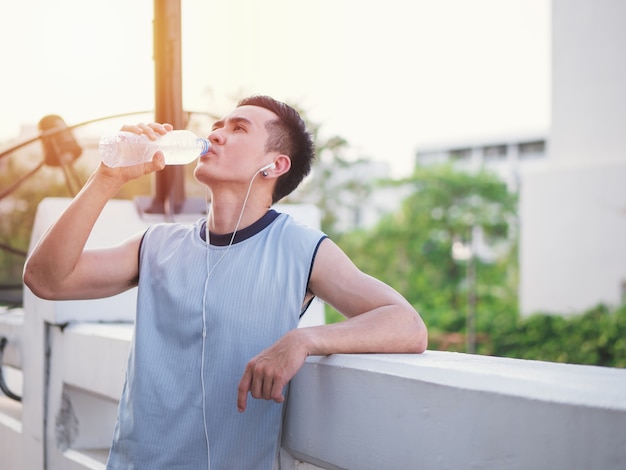 Giovane asiatico bello che ascolta la musica sugli earbuds e sull&#39;acqua della bevanda, stile di vita sano