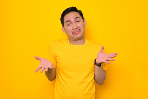Giovane asiatico confuso in maglietta casual che diffonde la mano reagendo a qualcosa di isolato su sfondo giallo Concetto di stile di vita delle persone