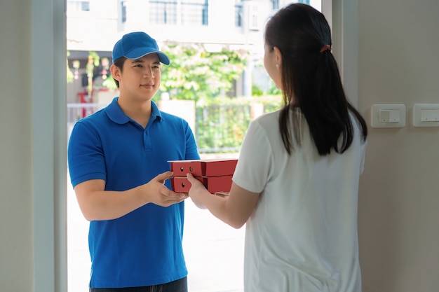 Giovane asiatico di consegna nel sorriso dell'uniforme blu e che tiene i contenitori di pizza in casa anteriore