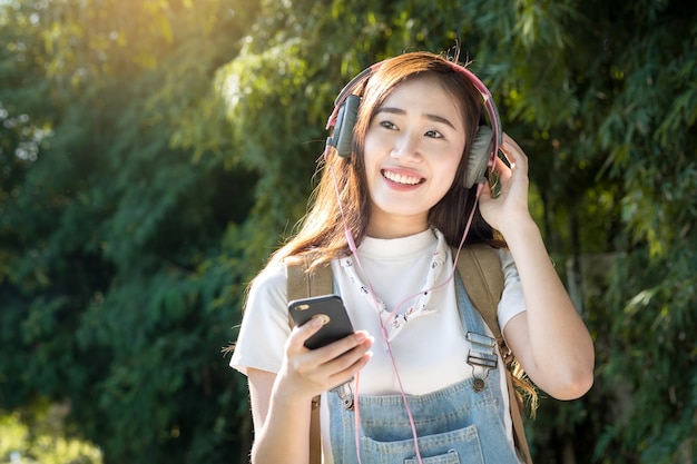 Giovane asiatico hipster indossa la cuffia che ascolta la musica attraverso smartphone in piedi all&#39;aperto sullo sfondo della natura