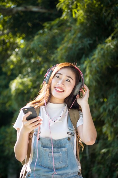 Giovane asiatico hipster indossa la cuffia che ascolta la musica attraverso smartphone in piedi all&#39;aperto sullo sfondo della natura