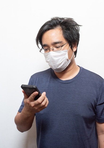 Giovane asiatico in casual che indossa una maschera medica chirurgica per proteggere COVID-19 utilizzando un telefono cellulare intelligente isolato su bianco Concetto di assistenza sanitaria. Prevenzione delle epidemie di Wu coronavirus (COVID-19) in pubblico ar