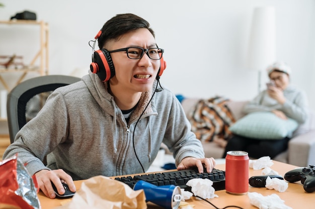 giovane asiatico in cuffia dedito ai giochi online per computer. ragazzo cinese concentrato che digita sulla tastiera seduto su una scrivania disordinata. la vista sfocata della ragazza si rilassa utilizzando il cellulare sul divano sul retro.